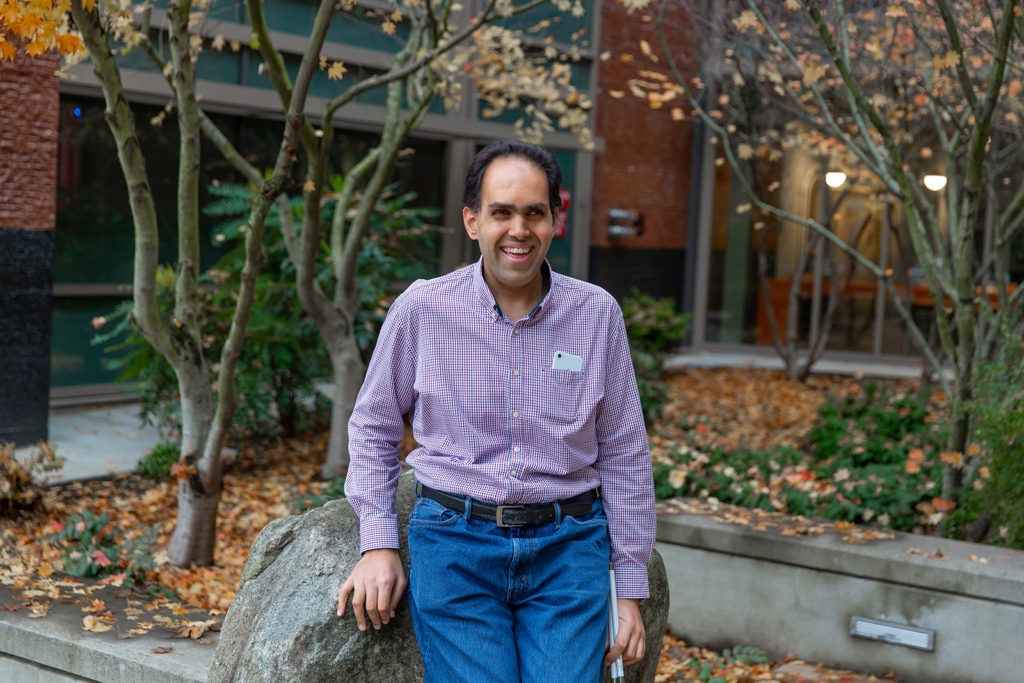 Microsoft’s Saqib Shaikh smiling and standing outside a Microsoft office building leaning against a rock with a backdrop of pretty Japanese maple trees shedding their golden leaves